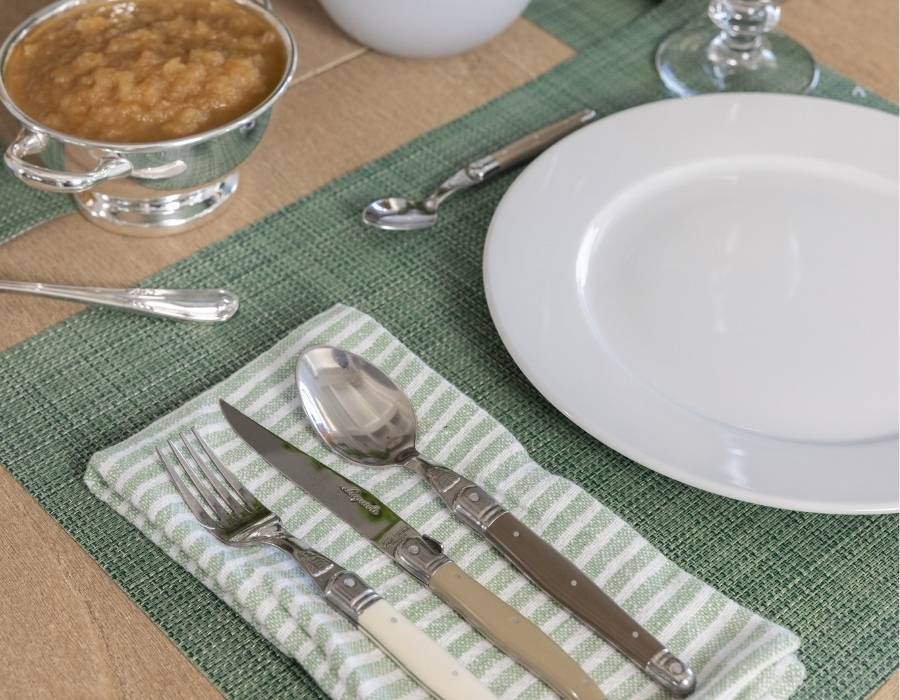 Elegant place setting with striped green napkin, vintage cutlery, white dinner plate, silver serving dish with applesauce, and woven green placemat on a wooden table.
