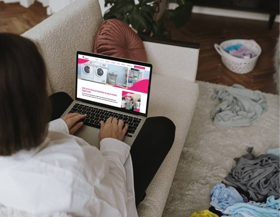 Woman sitting on couch using laptop to book professional laundry or cleaning services online, surrounded by unfolded laundry and laundry baskets in cozy home setting.