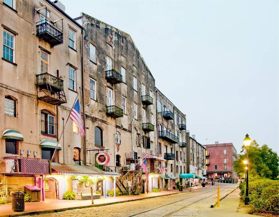 Historic riverfront buildings with iron balconies and cobblestone streets along River Street in Savannah, Georgia, featuring shops, American flags, and glowing street lamps at dusk.