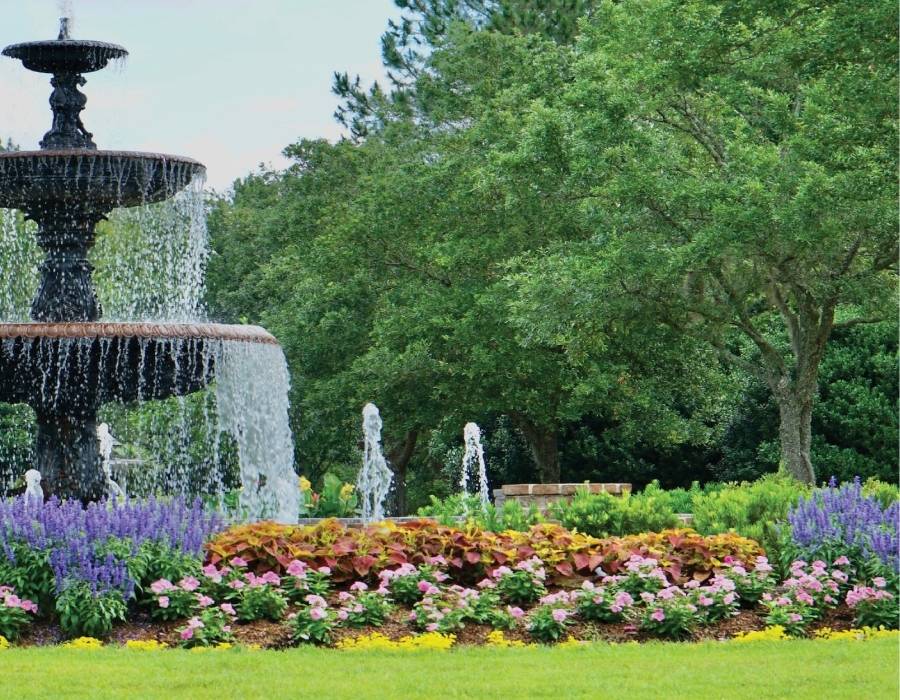 Beautiful landscaped garden featuring a tiered water fountain, colorful flower beds with seasonal blooms, and lush green trees in a professionally maintained public park.